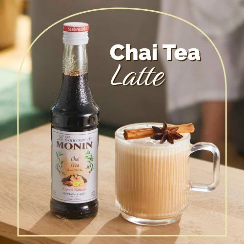Monin Chai Tea syrup bottle next to a chai tea latte with cinnamon sticks on a wooden surface.