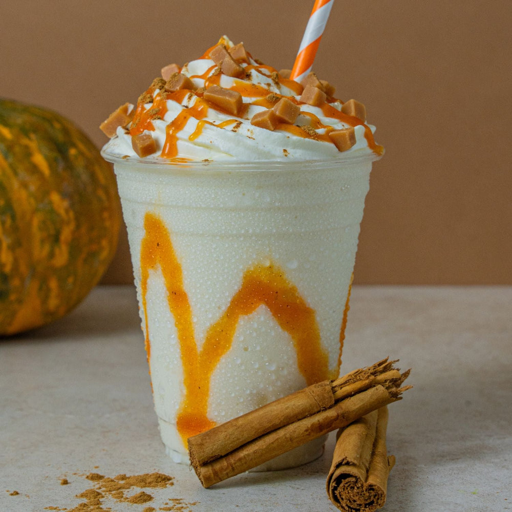 A frosted iced coffee or milkshake in a clear plastic cup, topped with whipped cream, caramel drizzle, and caramel pieces, with an orange-and-white striped straw. Cinnamon sticks and a pumpkin are visible in the background and foreground against a warm brown backdrop.