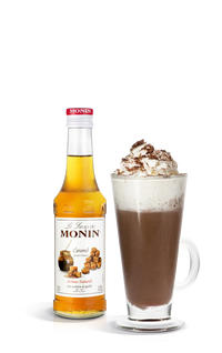Monin caramel syrup bottle next to a glass of hot chocolate with whipped cream on a white background