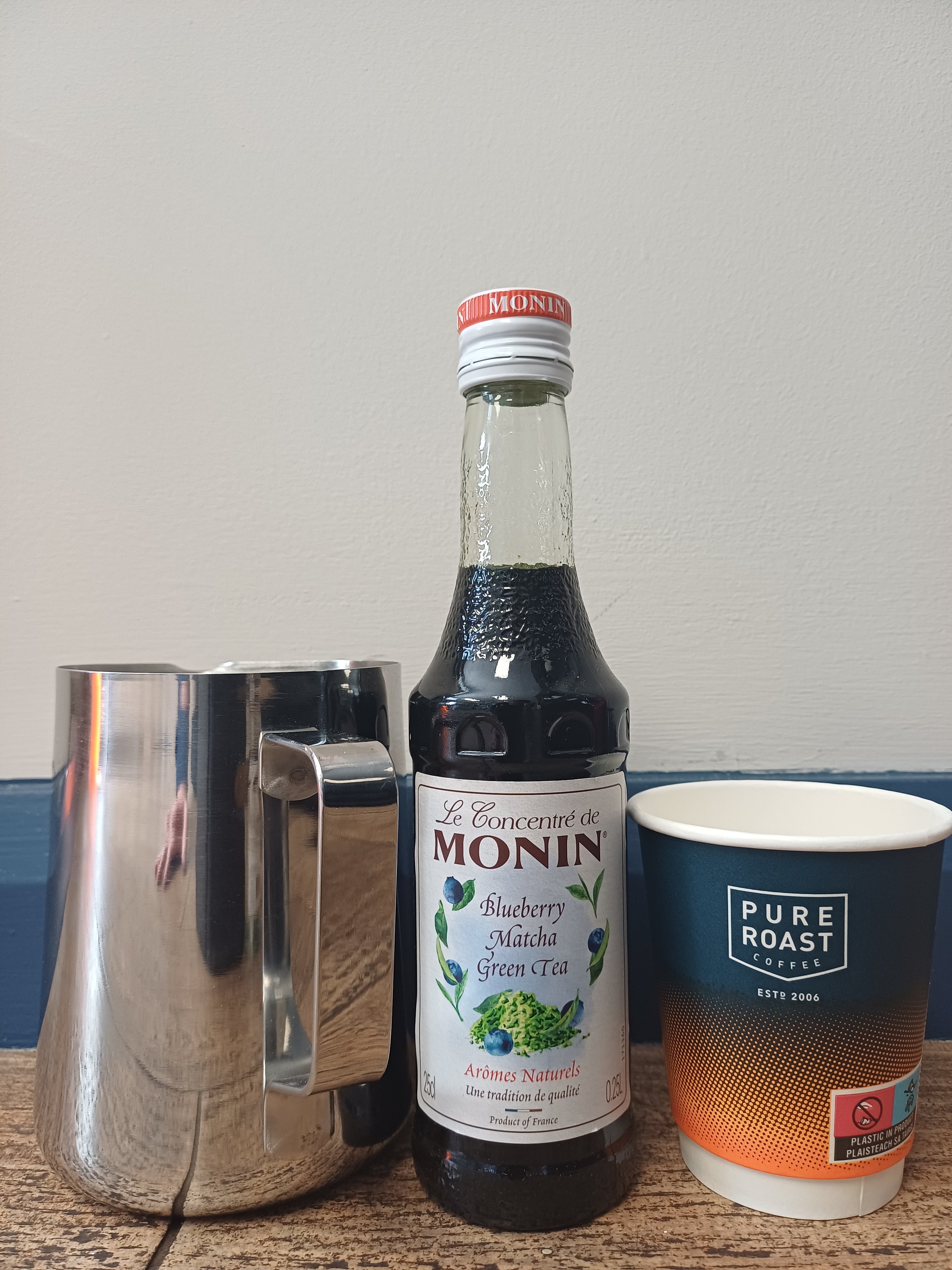 Monin_blueberry_matcha_green_tea_concentrate_bottle_product_setup_with_stainless_steel_pitcher_and_paper_cup