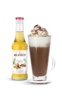 Monin gingerbread syrup bottle next to a glass of hot chocolate with whipped cream on a white background