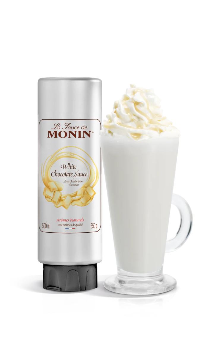 A bottle of Monin White Chocolate Sauce next to a glass of coffee topped with whipped cream.