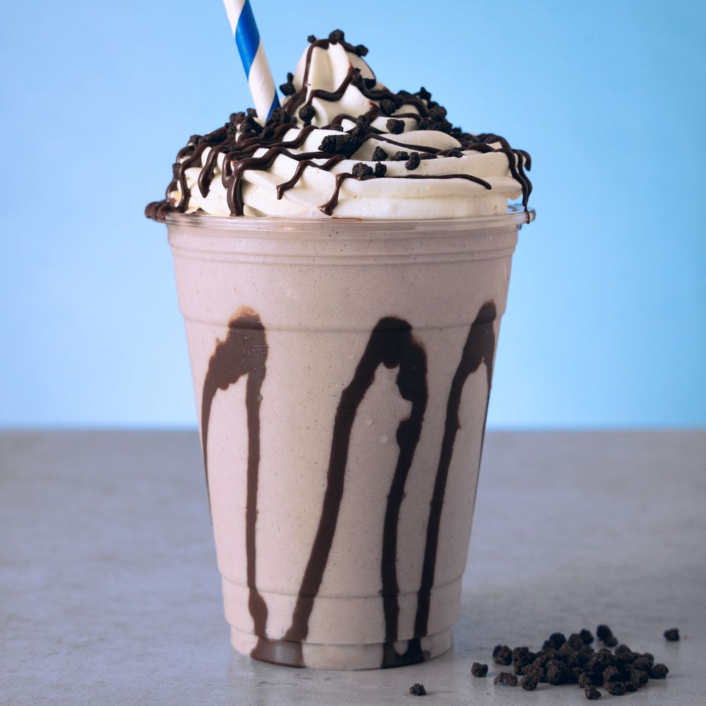 A chocolate milkshake in a clear plastic cup with whipped cream, chocolate drizzle, and chocolate sprinkles on top, with a blue-and-white striped straw, set against a light blue background.