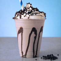A chocolate milkshake in a clear plastic cup with whipped cream, chocolate drizzle, and chocolate sprinkles on top, with a blue-and-white striped straw, set against a light blue background.