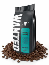 A black coffee bag standing upright on a white background, surrounded by scattered roasted coffee beans. The bag has large distressed white vertical text reading 'WANTED' and a teal label that says 'Colombian Supremo' and 'Whole Bean'.