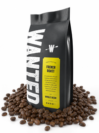 A black bag of WANTED French Roast whole bean coffee stands upright on a white background, surrounded by scattered roasted coffee beans. The bag features a large distressed white 'WANTED' logo and a bright yellow label with black text.