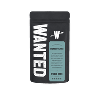 A matte black coffee bag with a large distressed white 'WANTED' logo on the left and a light blue-green label on the front reading 'Fresh Roasted Metropolitan Whole Bean'.