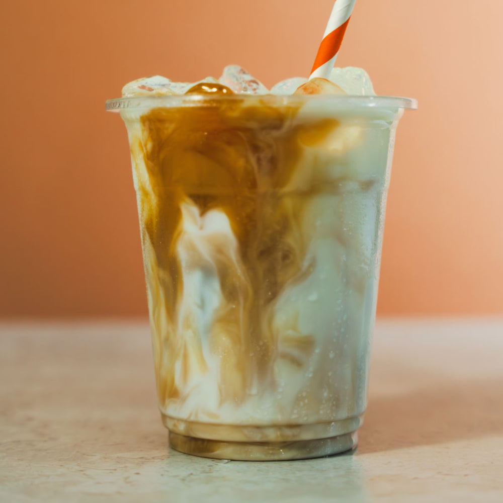 An iced coffee drink in a clear plastic cup with a striped paper straw, showing swirls of white milk and dark coffee with caramel drizzle, set against a warm peach background.