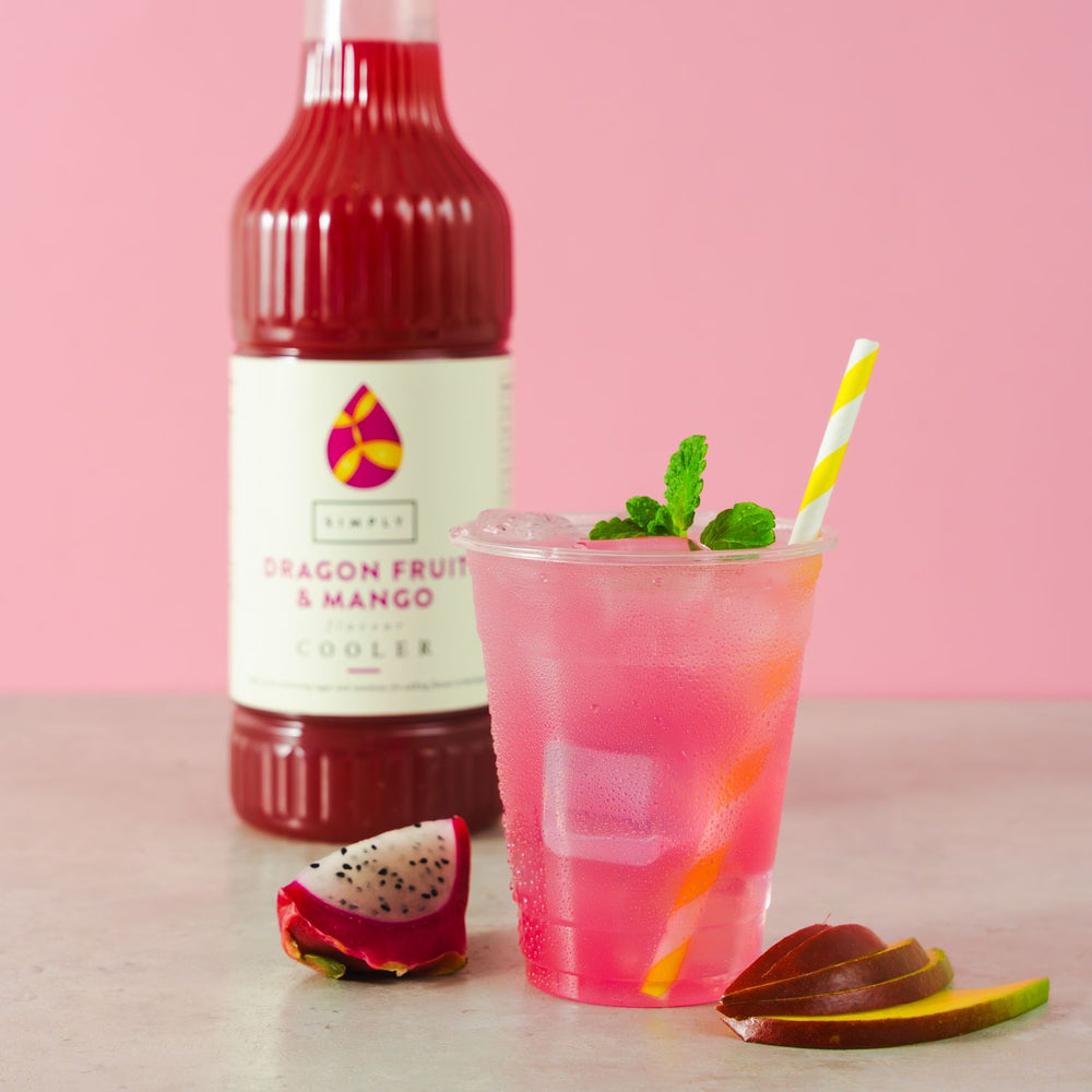 A pink dragon fruit and mango cooler drink in a clear plastic cup with a yellow-and-white striped straw, set on a light surface with a pink background. A bottle of Simply Dragon Fruit & Mango Cooler stands behind the drink, with dragon fruit and mango slices nearby.