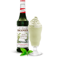 A glass bottle of Monin Matcha Green Tea syrup next to a glass filled with a green beverage and ice, garnished with a mint leaf.