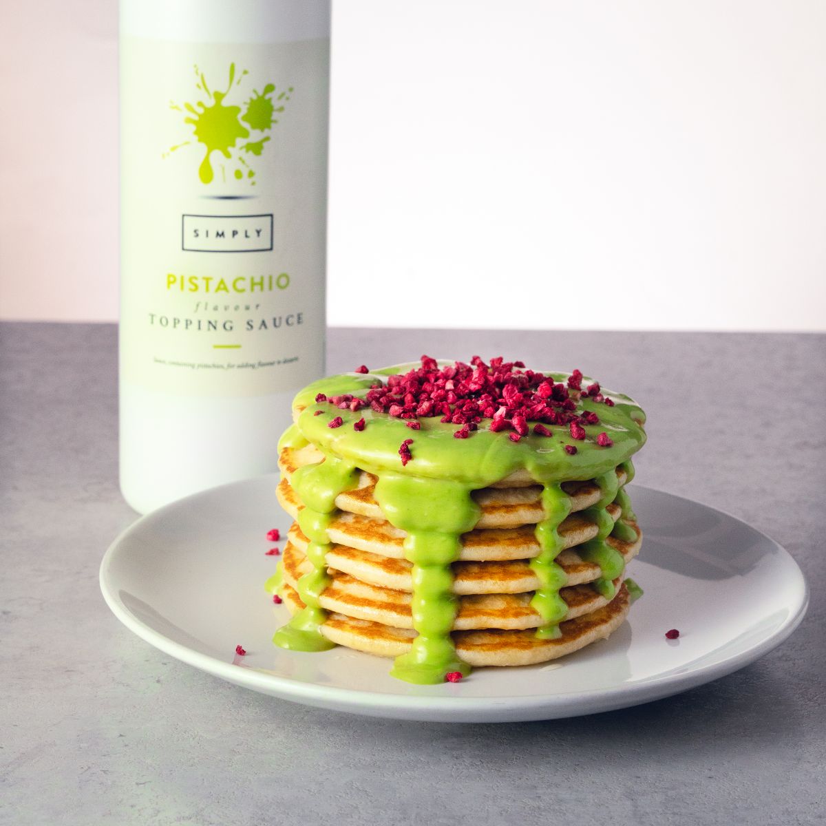 A stack of pancakes topped with bright green pistachio-flavored topping sauce and pink sprinkles, served on a white plate with a bottle of Simply Pistachio topping sauce in the background.