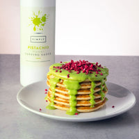 A stack of pancakes topped with bright green pistachio-flavored topping sauce and pink sprinkles, served on a white plate with a bottle of Simply Pistachio topping sauce in the background.
