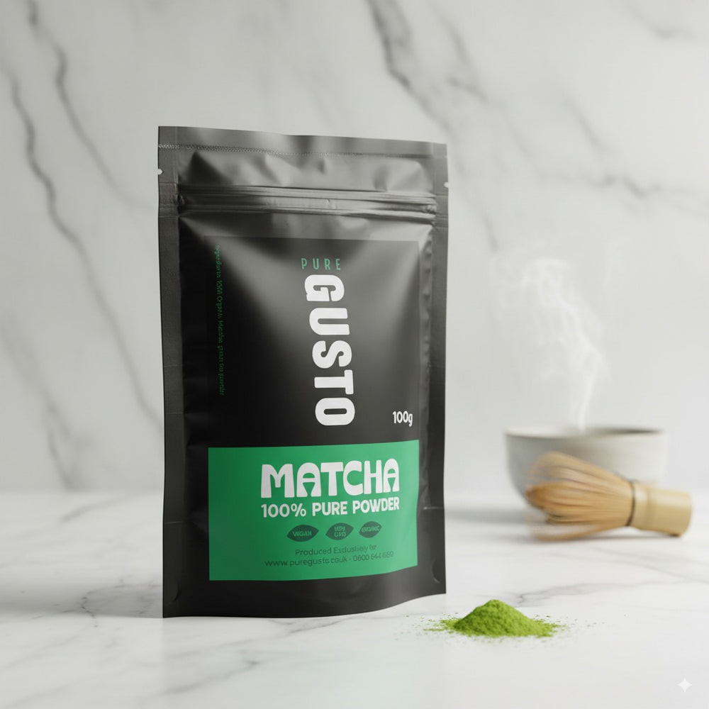 Pure Gusto 100g pouch of 100% pure organic matcha powder, displayed with a bamboo whisk and a small pile of vibrant green matcha powder, suggesting freshness and preparation, on a clean marble background.