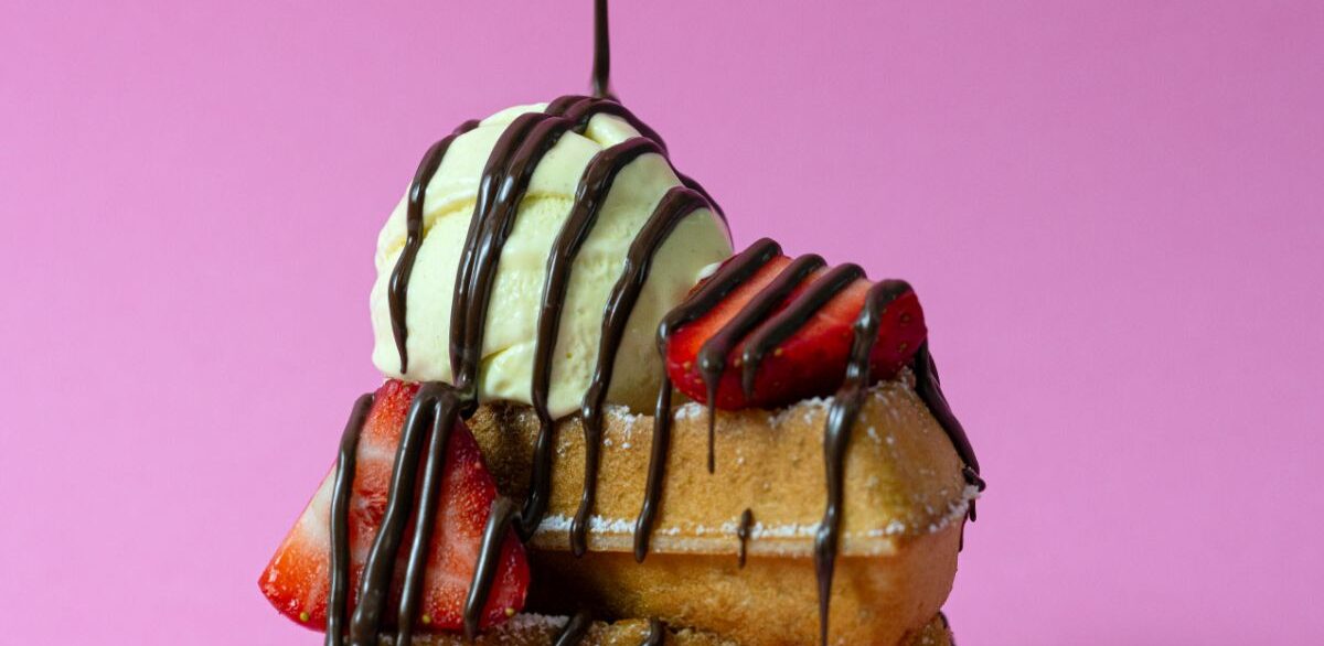 A close-up of a dessert stack on a pink background, featuring a waffle topped with a scoop of vanilla ice cream, fresh strawberry slices, and drizzles of chocolate sauce.