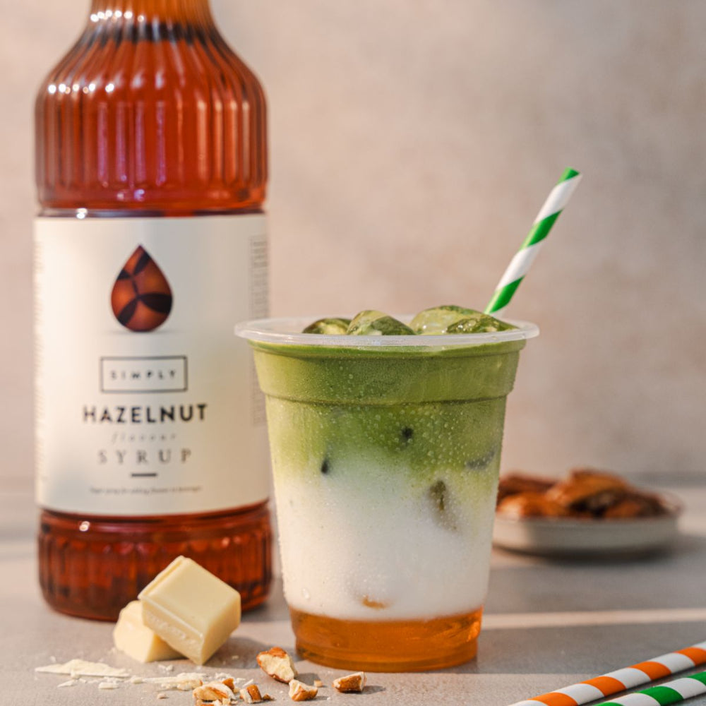 A clear plastic cup filled with a layered green and white iced drink and a green-and-white striped straw, placed in front of a bottle of Simply Hazelnut Syrup. White chocolate pieces and nut crumbs are scattered on the surface, with a softly blurred neutral background.