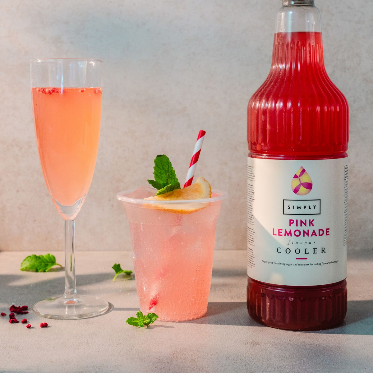 A bottle of Simply Pink Lemonade Refresh Cooler stands next to a filled champagne flute and a pink drink in a clear cup with a red-and-white striped straw, set on a light stone surface against a pale wall.