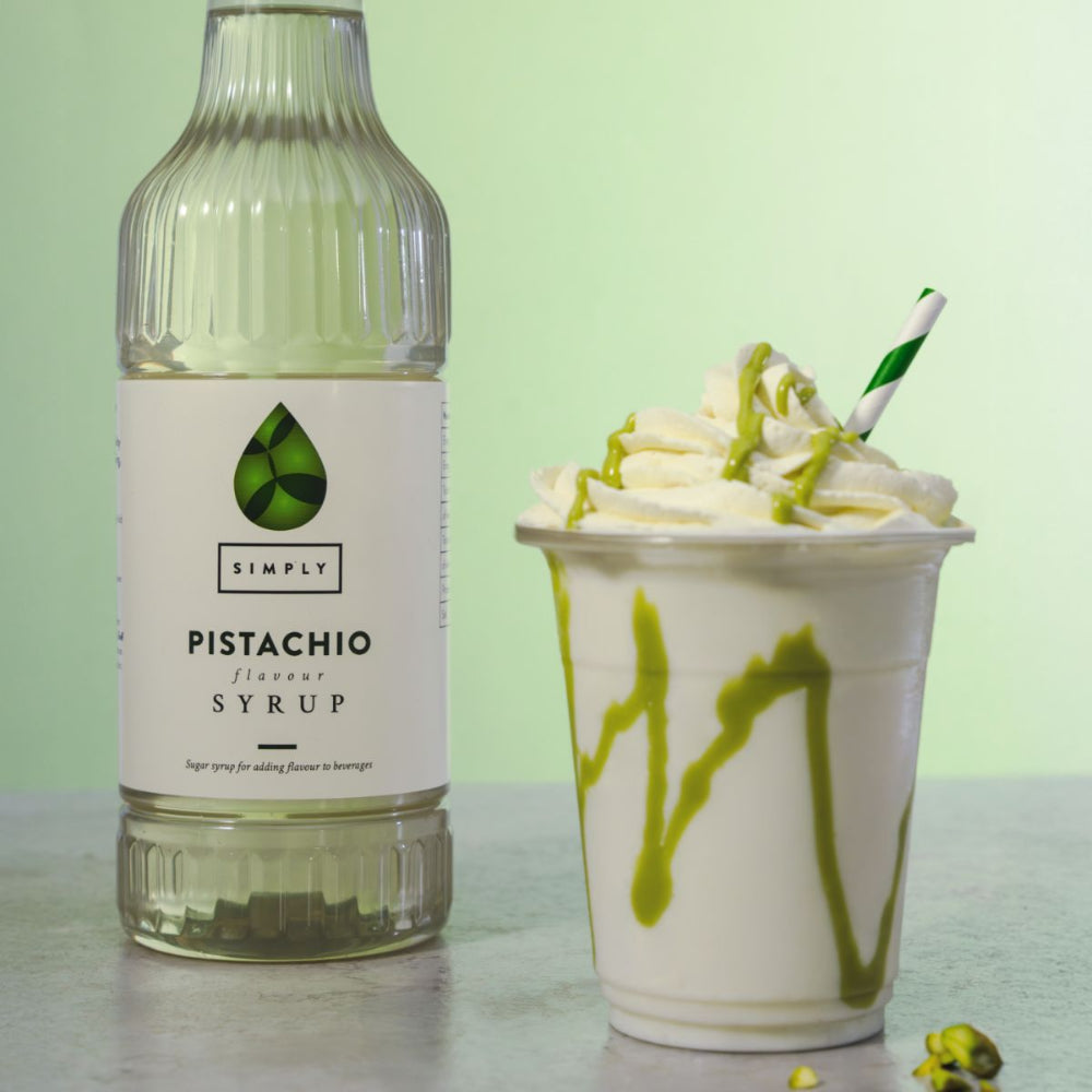 A bottle of Simply Pistachio Flavor Syrup stands next to a whipped cream drink topped with green syrup drizzle and a green-and-white striped straw, on a light surface against a pale green background.