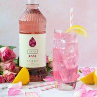 A bottle of Simply Rose flower syrup stands beside a tall glass of pink iced drink with a lemon wedge and striped straw, surrounded by pink roses, lemon slices, and pink petals on a light surface against a soft pink background.
