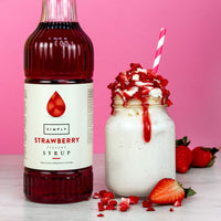 A bottle of Simply Strawberry Syrup stands next to a milkshake in a mason jar topped with whipped cream, strawberry drizzle, and a pink-and-white striped straw, set against a pink background with strawberries around it.