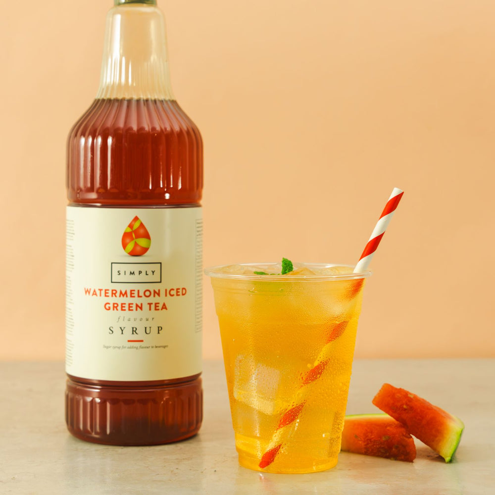 A bottle of Simply Watermelon Iced Green Tea Syrup stands next to a clear plastic cup filled with an orange iced drink and a red-and-white striped straw, with watermelon slices on a beige surface against a peach background.