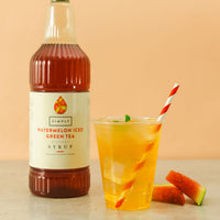 A bottle of Simply Watermelon Iced Green Tea Syrup stands next to a clear plastic cup filled with an orange iced drink and a red-and-white striped straw, with watermelon slices on a beige surface against a peach background.