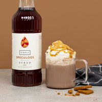 A bottle of Simply Speculoos flavor syrup stands next to a glass mug filled with a hot chocolate or cocoa drink topped with whipped cream, caramel drizzle, and cookie crumbs, with cookie pieces and a folded dark cloth on a neutral surface against a warm tan background.