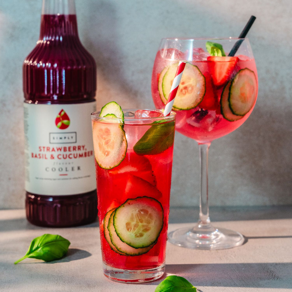 A bottle of Simply Strawberry Basil & cucumber Cooler drink stands behind two prepared cocktails: a short glass with red drink, cucumber slices, basil leaves, and a striped straw, and a larger wine glass with pink drink, cucumber slices, strawberries, and a black straw. Styled on a light surface against a gray wall.