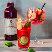 A bottle of Simply Strawberry Basil & cucumber Cooler drink stands behind two prepared cocktails: a short glass with red drink, cucumber slices, basil leaves, and a striped straw, and a larger wine glass with pink drink, cucumber slices, strawberries, and a black straw. Styled on a light surface against a gray wall.